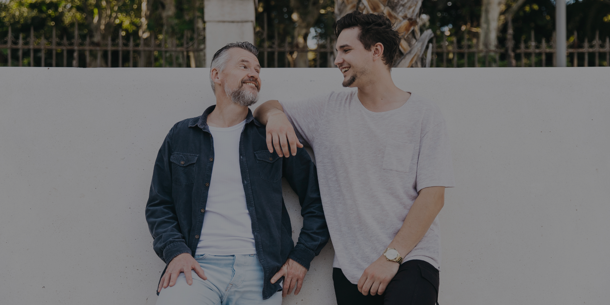 Two men standing close together against a white wall with trees in the background

