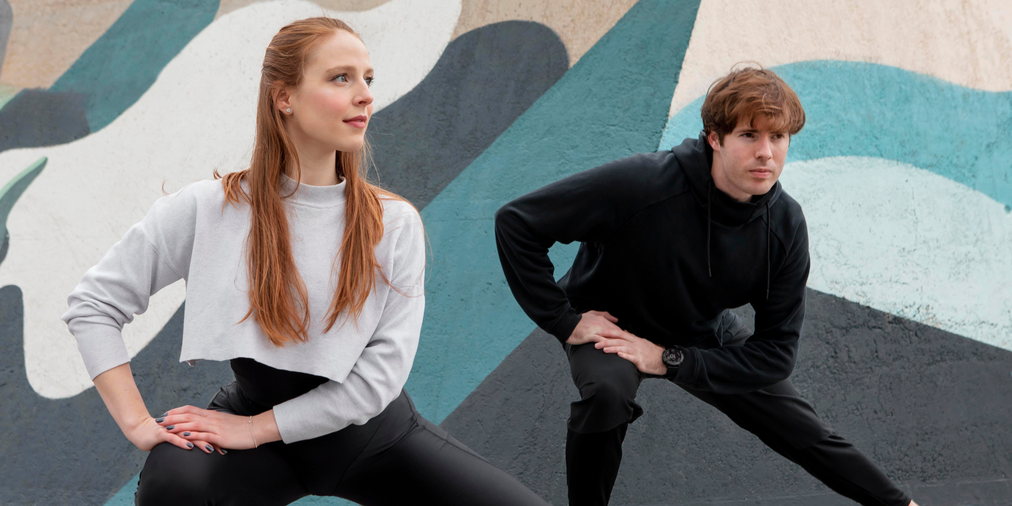Two adults warming up outdoors, stretching in front of a colorful wall.