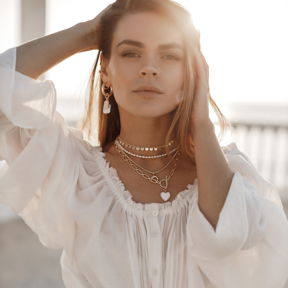 Woman wearing a white blouse and multiple necklaces with a blurred background

